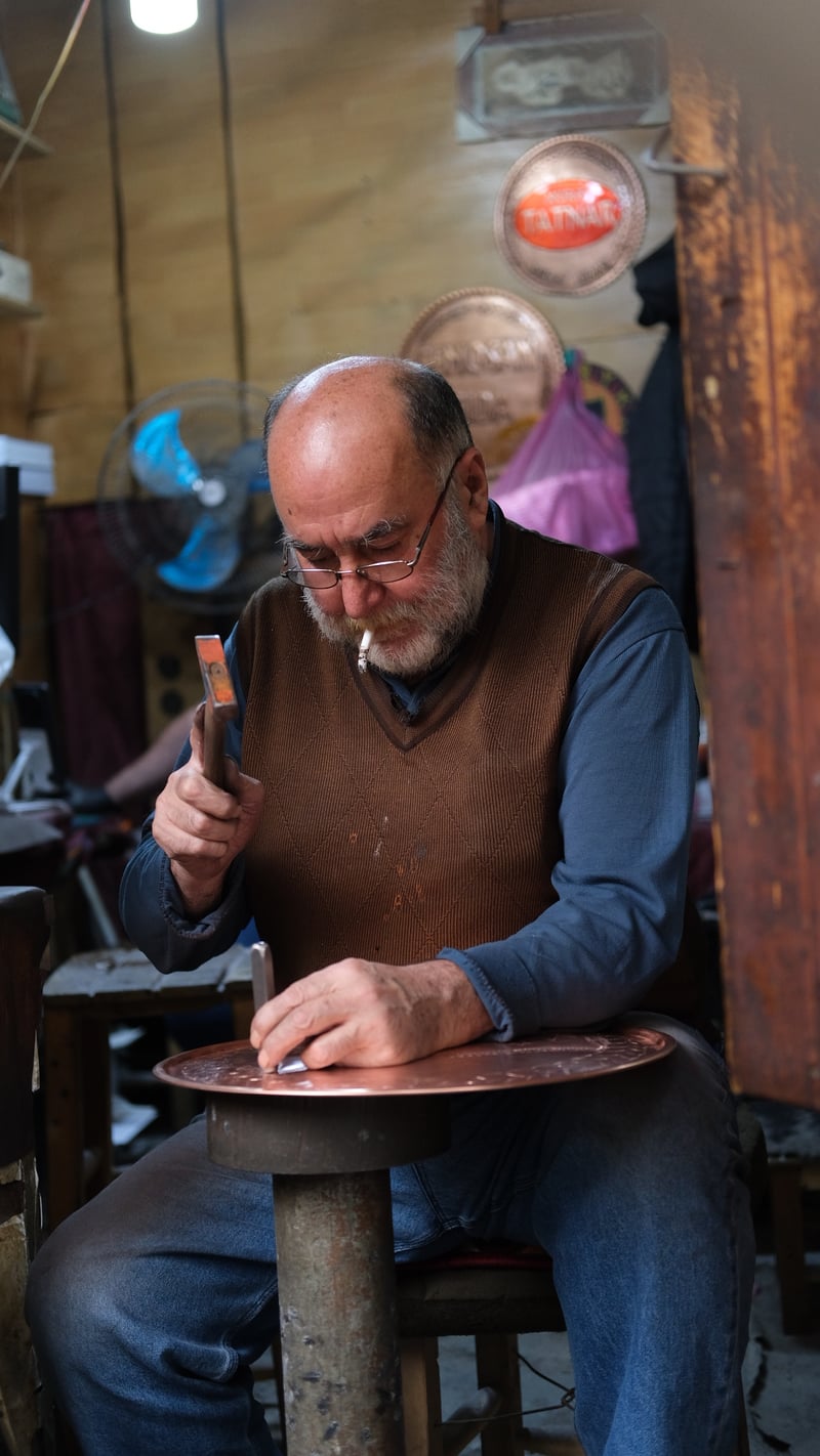 an old coppersmith with a cigarette hammering a pattern into a copper tray in his workshop