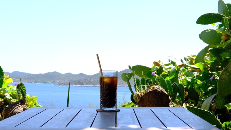 a glass of iced coffee on a wooden table, cactus and a blue bay behind