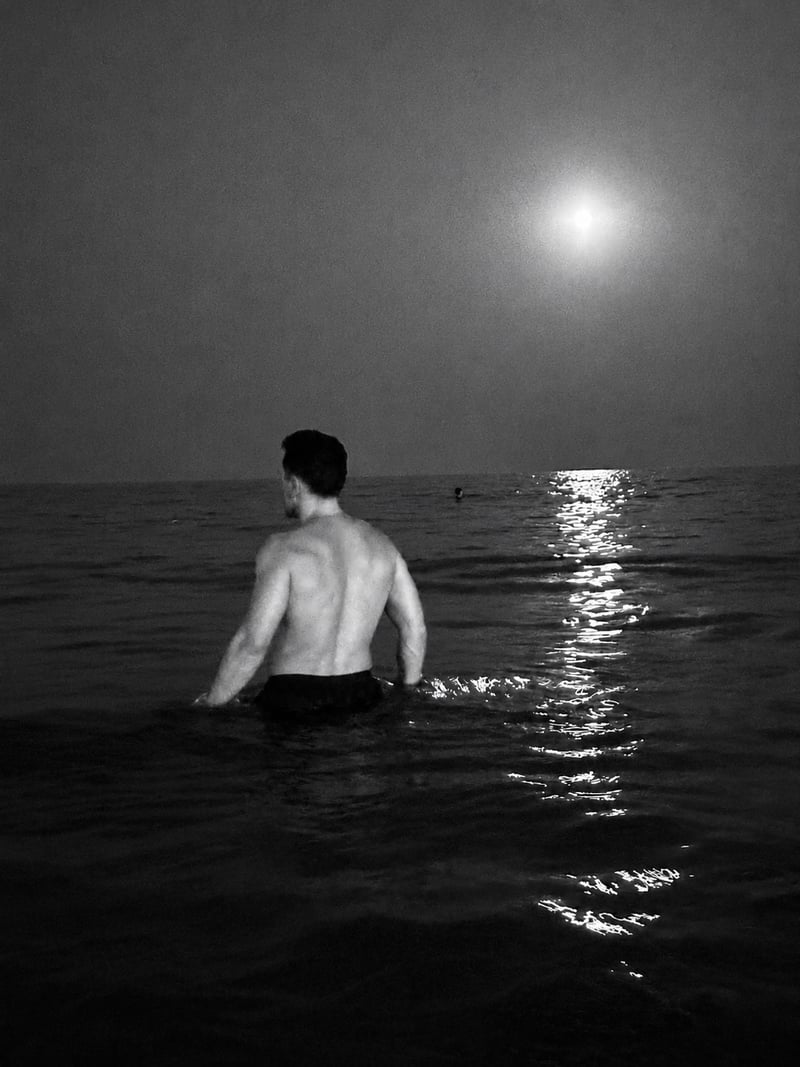 a man standing waist deep in a dark sea, looking at the moon and its path of light on the water