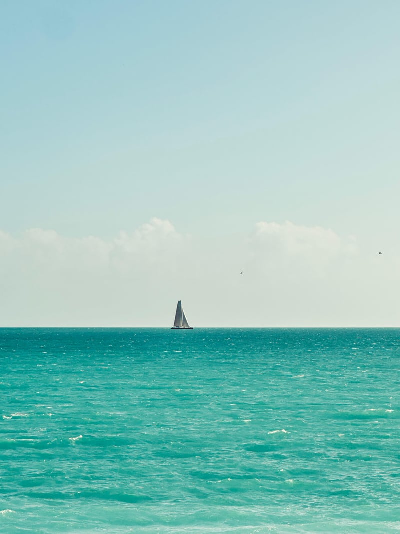 a white sailboat on the horizon between a turquoise sea and a pale blue sky